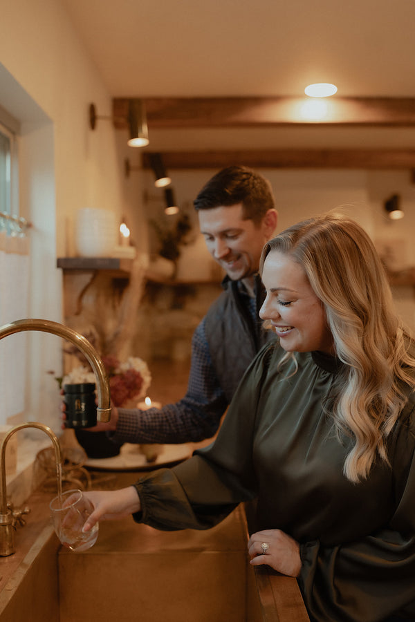 Couple smiling in a kitchen, man reaching for a shelf item while woman fills a glass of water at the sink
